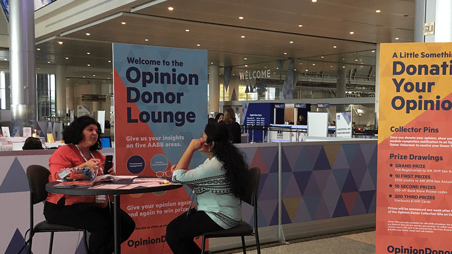 Picture of 2 women talking at a table in front of a sign reading "Welcome to the Opinion Donor Lounge"