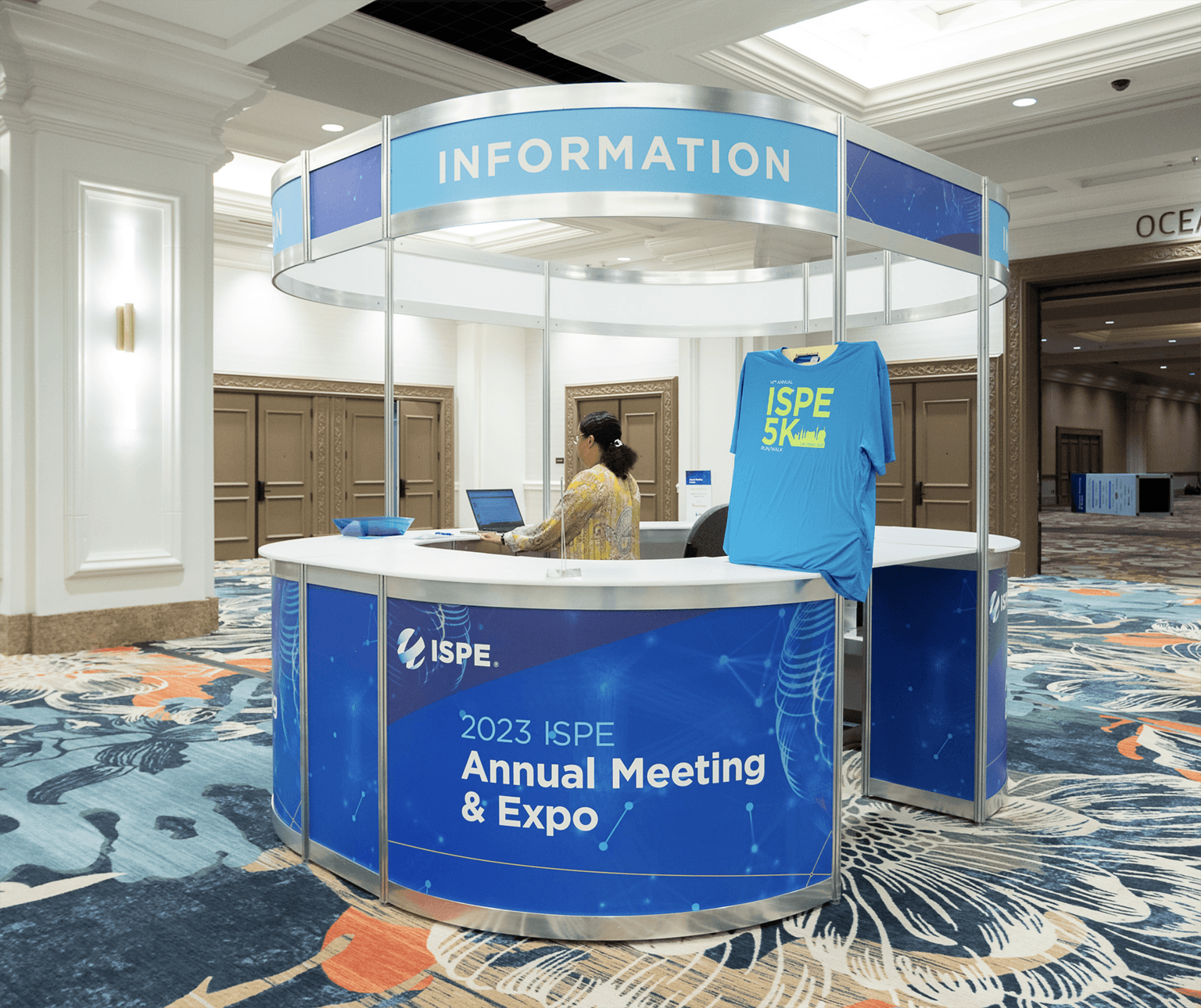 Circular information booth for the 2023 ISPE Annual Meeting & Expo. The booth is covered in blue ISPE branding, with an “Information” banner above and an ISPE 5K T-shirt displayed on the front.